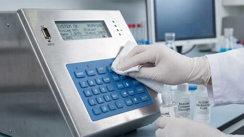A technician in white gloves wipes a blue silicone keypad on a laboratory device using a cloth and specialized cleaning liquid