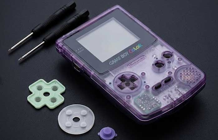 A clear purple Game Boy Color console displayed on a black surface alongside two screwdrivers and several replacement silicone button pads and conductive rubbers