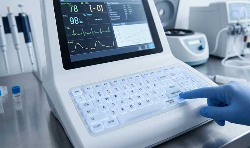 A gloved hand operating a medical diagnostic device featuring integrated backlit silicone keyboards with a sealed cleanable surface and soft white illumination in a laboratory setting