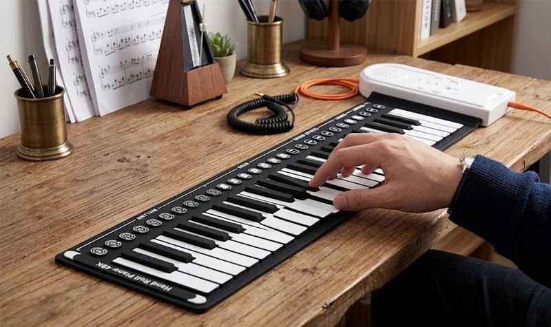A hand pressing a key on a black and white Silicone Piano Keyboard placed flat on a wooden desk with sheet music