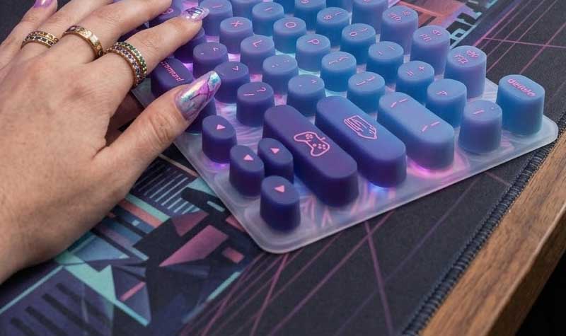 A hand with decorated long nails types on a vibrant vaporwave themed silicone keyboard for long nails at a matching gaming setup