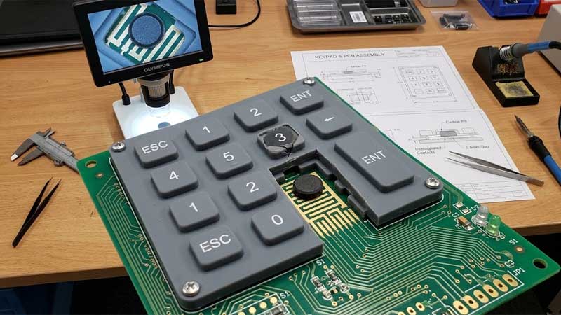 An engineering inspection setup shows carbon pills under a silicone keypad aligned with gold PCB contact patterns on a workbench