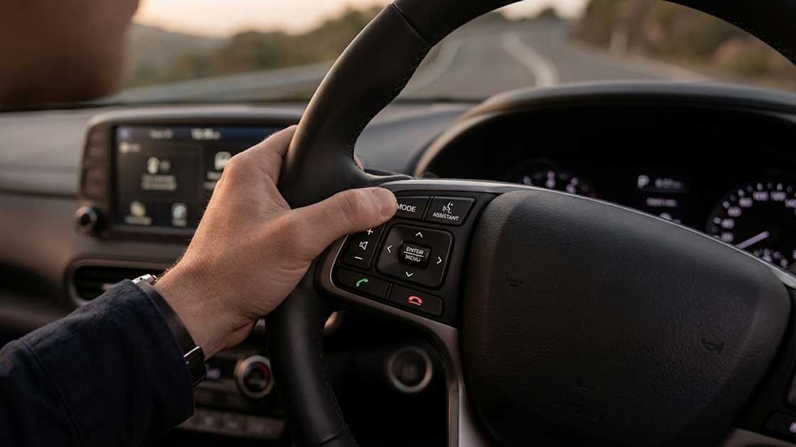 A driver uses their thumb on the automotive electronics silicone keypads on a steering wheel while driving at sunset