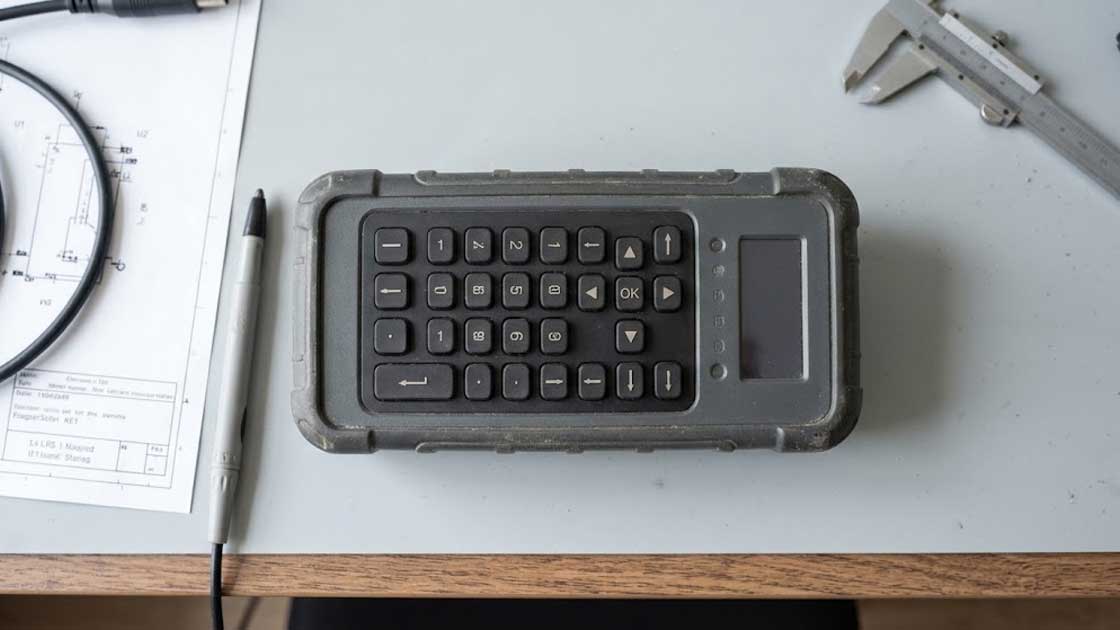 A handheld electronic device with a conductive silicone keypad lies flat on a technical workbench alongside tools and schematics