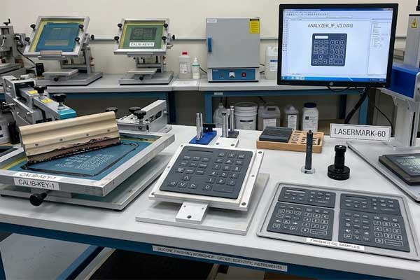 Printed laboratory equipment silicone keypad samples rest on a workshop desk between screen printing and laser marking tools