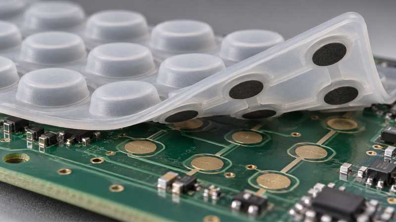 A translucent silicone keypad with black conductive contacts is lifted above a circuit board